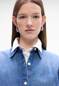 Camicia in denim di un blu chiaro, con una camicia bianca a collare sotto, e orecchini rotondi in argento. Chiusura con bottoni e dettagli di cucitura discreti.