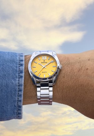 Montre en métal argenté avec un cadran jaune comportant le chiffre romain XII, un mouvement à quartz et un fermoir à bracelet poli, portée au poignet.