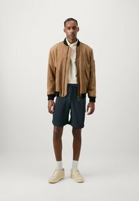 Giacca bomber in suede marrone con dettagli neri, maglione a maglia crema, pantaloni corti blu navy e sneakers beige. Modella in piedi contro uno sfondo neutro.