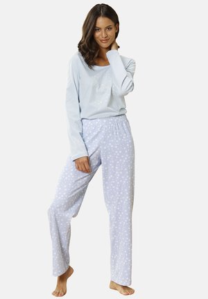 Frau trägt ein hellblaues Langarmshirt und passende Schlafanzughose mit Sternenmuster, steht barfuß vor einem schlichten Hintergrund.