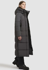 Cappotto lungo imbottito di un grigio scuro con colletto alto e chiusura con zip. Presenta una trapuntatura rettangolare e tasche laterali con zipper. Materiale pesante e isolante.