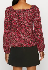 Blouse rouge à fleurs avec un col carré, des manches longues bouffantes et un dos froncé. Associée à une jupe noire évasée dotée d'une fermeture éclair.