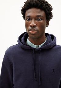 Hemme bleu marine avec cordon de serrage, arborant un petit logo noir sur la poitrine. Porté par-dessus une chemise à col bleu clair. Texture de tissu douce.