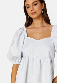 Světle modré šaty s vypufovanými krátkými rukávy, čtvercovým výstřihem a texturovanými květinovými vzory po celém povrchu. Mají nařasený pas.