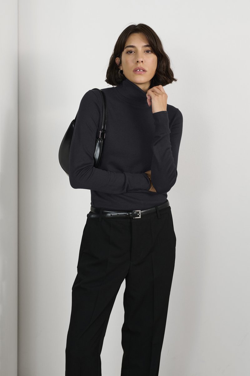 Un haut col roulé noir en matière côtelée, associé à un pantalon noir taille haute. Le mannequin porte un petit sac noir.