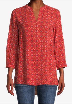 Frau trägt rote Tunika mit orange-lila kreisförmigen Blumenmustern, einem V-Ausschnitt und dreiviertellangen Ärmeln, kombiniert mit blauer Jeans.