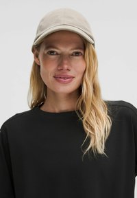 Casquette beige clair avec visière incurvée et logo brodé ; portée sur un t-shirt noir à col rond. Les cheveux sont longs et ondulés, encadrant le visage.