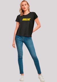 Black t-shirt with yellow "BILLIE" text, short sleeves, relaxed fit. Paired with blue skinny jeans and white sneakers.
