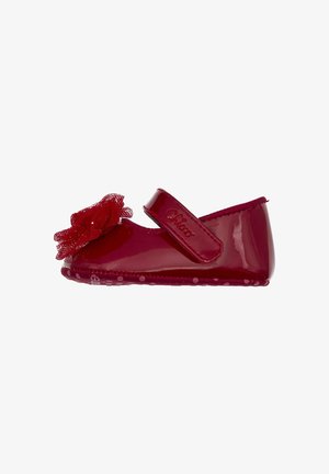 Chaussure en cuir verni rouge avec une bride, ornée d'une fleur en maille sur le bout, d'une semelle à pois et d'une texture lisse.