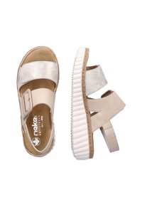 Beige sandaler med metalliskt silverfärgade remmar, elastiska band och en vit, texturerad sula. Har en justerbar spänne och vadderad innersula.