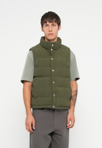 Jachetă pufosă verde măsliniu cu guler înalt, material texturat, două rânduri de nasturi metalici argintii, purtată peste o cămașă cu mânecă scurtă de culoare gri deschis.
