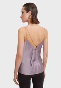 Levandulový saténový top na ramínka s tenkými řetízky, vyznačující se klíčovou dírou na zádech a volným střihem, v kombinaci s černými kalhotami.