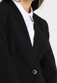 Kvinna som bär stora hoopörhängen, vit skjorta med krage och en svart kavaj med en enkel knapp knäppt.