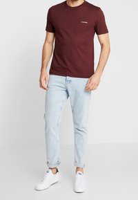 Bordeaux katoenen t-shirt met ronde hals en logo, gecombineerd met lichtblauwe opgerolde jeans en witte sneakers met marineblauwe accenten.