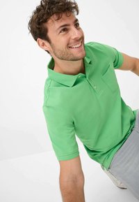 Polo verde in cotone con colletto, maniche corte e taschino sul petto. Modello indossa pantaloni grigi chiari e scarpe.
