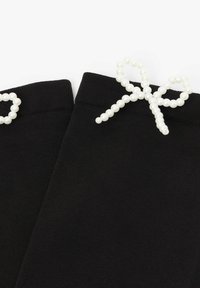 Czarne skarpetki o gładkiej fakturze, z białą kokardką zdobioną perłowymi koralikami blisko góry. Projekt jest prosty i elegancki.