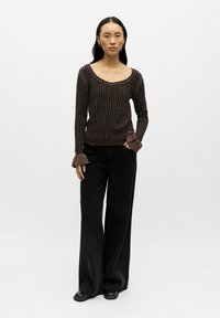 Kvinde står foran en hvid baggrund iført en sort og brun ternet sweater med flagrende ærmer, brede sorte bukser og sorte sko.