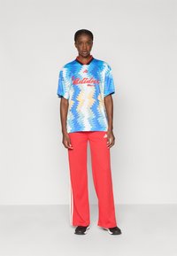 Kobieta stoi na jednolitym tle, ubrana w niebieską koszulę Adidas w wzory oraz czerwone spodnie dresowe Adidas z białymi paskami.