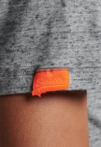 Tessuto di maglietta grigia con un motivo testurizzato; presenta un'etichetta arancione vivace con il testo "SuperDry" e un design a bordo distintivo.