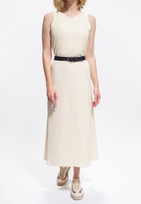 Donna che indossa un abito midi beige senza maniche con cintura intrecciata nera e sneakers beige con dettagli a stampa leopardata, in piedi davanti a uno sfondo bianco.