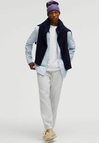 Mandlig model iført en lyseblå stribet skjorte, hvid t-shirt, navy-farvet ærmeløs sweater, grå sweatpants og beige sko samt en lilla hue.