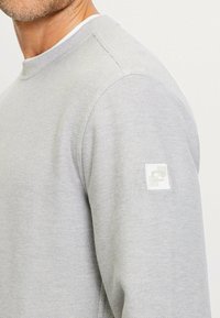 Hellgraues Sweatshirt mit strukturierter Oberfläche, ausgestattet mit einem gerippten Kragen und einem kleinen weißen Logopatch an Ärmelsaum links.