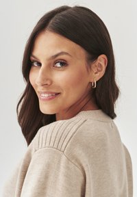 Beige trui met gebreide schouderdetails, gedragen met kleine gouden oorbellen, afgebeeld tegen een effen lichte achtergrond.