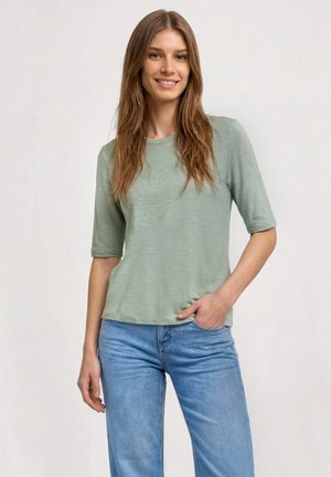 Femme aux longs cheveux bruns portant un t-shirt vert clair à manches courtes et un jean bleu, souriante avec une main dans sa poche.