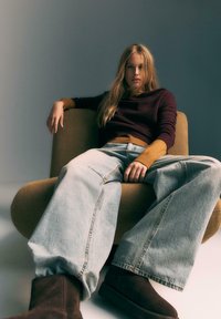 Młoda kobieta z długimi blond włosami nosi oversize'owe jasne dżinsy, ciemne buty i warstwowy sweter, siedząc swobodnie na musztardowym fotelu.
