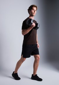 Svart hantel holdt i høyre hånd, båret av en mannlig figur i en mørkebrun t-skjorte og svarte shorts, som står foran en nøytral bakgrunn.