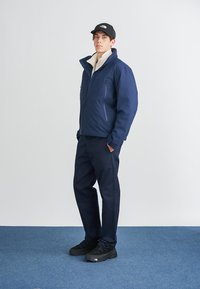 Marineblå isolert jakke med ståkrage, to glidelåslommer, kremfarget foring, brukt med mørke bukser og svarte joggesko.