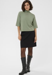 Zöld, rövid ujjú garbó pulóver, fekete ceruzaszoknyával és térdig érő fekete csizmával párosítva. Sima anyag, minimalista kialakítás.