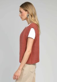 Gilet tricoté de couleur rouille, avec une coupe carrée, des manches courtes et des fentes latérales, superposé sur un T-shirt blanc. Texture lisse.