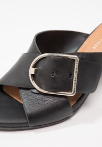 Sandale en cuir noir avec de larges brides croisées et une boucle en métal argenté gravée « Tommy Hilfiger » sur fond blanc.