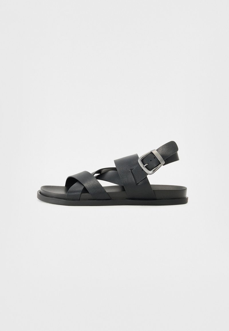Svart lær sandal med kryssende stropper og en justerbar spenne på hælen, med flat såle og minimalistisk design.