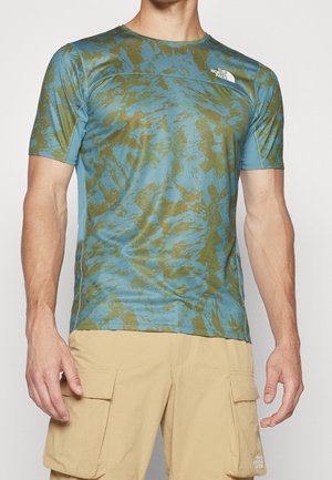 Homme portant un t-shirt à manches courtes bleu et olive à motif et un short cargo beige, debout devant un fond uni.