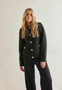 Musta teksturoitu takki, jossa on kultaiset napit ja kaksi etutaskua, yhdistetty mustiin housuihin. Minimalistinen muotoilu, jossa on istuva siluetti.