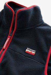 Tamsiai mėlynas vilnos striukė su raudonomis apdailomis ir užtrauktuku. Yra mažas išsiuvinėtas „SUPERDRY“ logotipas ir kalnų motyvas pleistre.