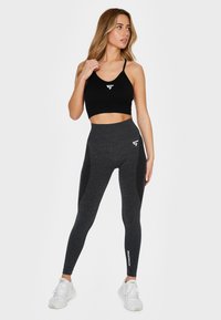 Black sports bra and high-waisted charcoal leggings with a textured pattern. White logo accents on the chest and thigh. White athletic shoes.