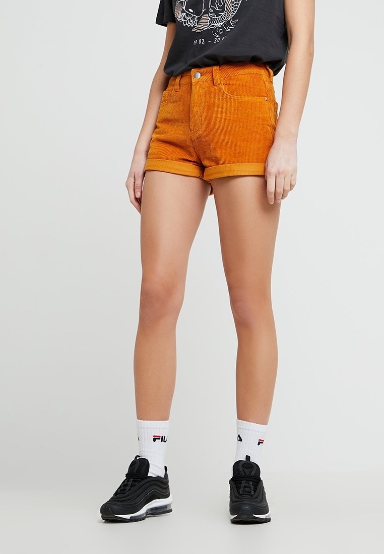 Personne portant un short en velours côtelé orange retroussé, un t-shirt noir à motif graphique, des chaussettes de sport blanches et des baskets noires, debout devant un fond uni.