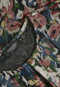 Bluse mit floralen Mustern aus mehrfarbigem Mesh-Stoff. Verfügt über einen runden Ausschnitt, kleinen braunen Knopfverschluss und ein Etikett "SOAKED IN LUXURY".
