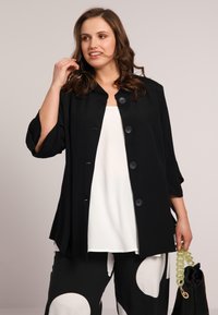 Veste légère noire avec trois boutons, coupe ample, et manches raccourcies, portée avec un haut blanc et un pantalon noir à motifs. Sac tendance inclus.