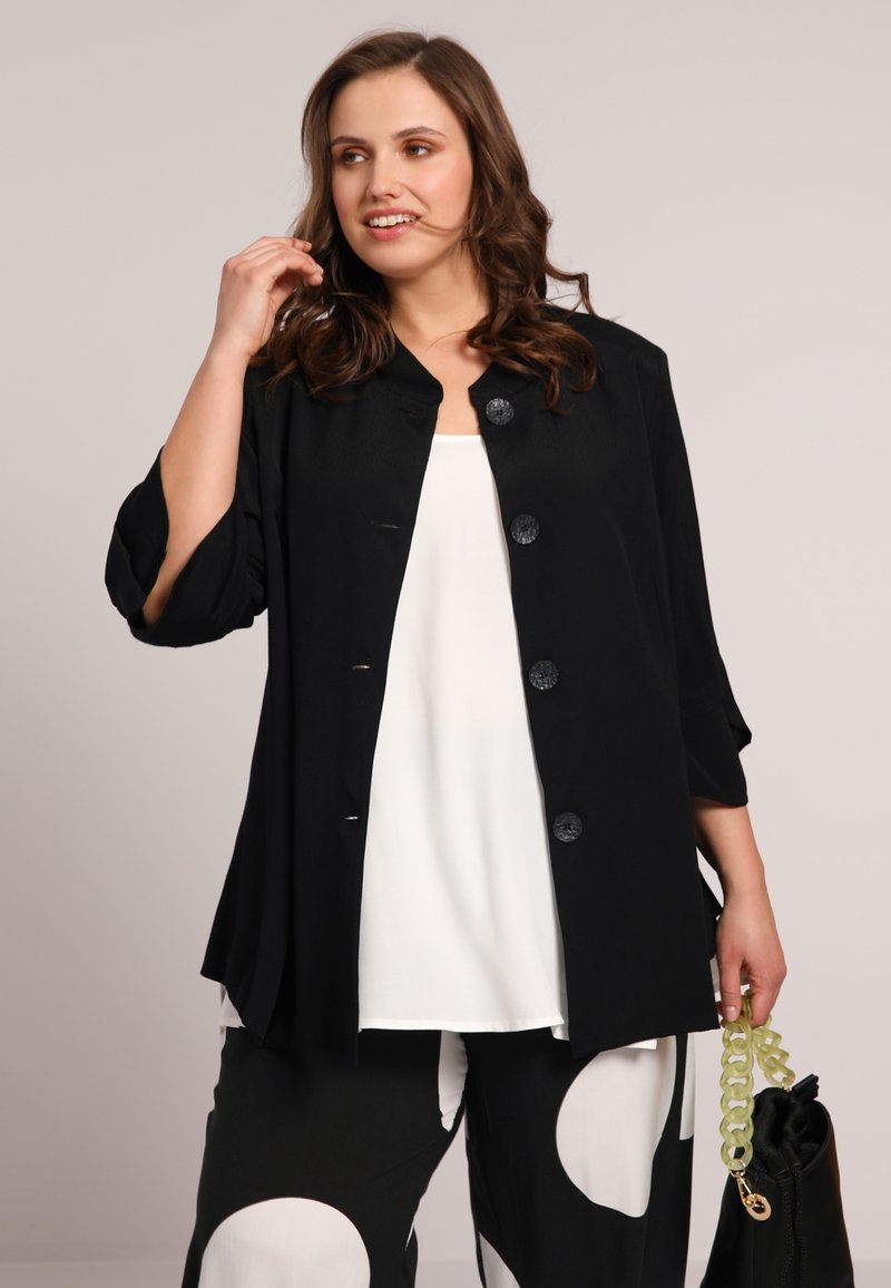 Veste légère noire avec trois boutons, coupe ample, et manches raccourcies, portée avec un haut blanc et un pantalon noir à motifs. Sac tendance inclus.