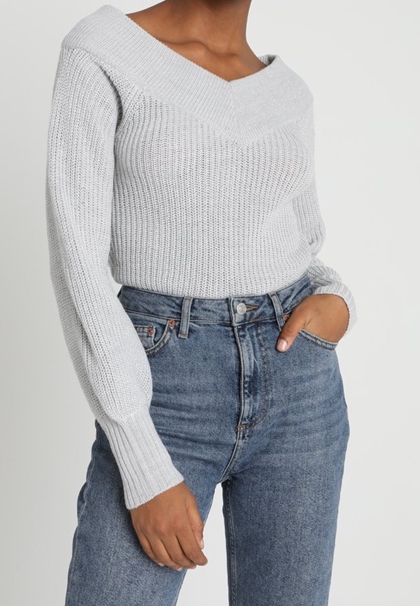 Světle šedý pletený svetr s odhalenými rameny a širokým výstřihem, s ribbovanou texturou a dlouhými rukávy, v kombinaci s džíny s vysokým pasem.