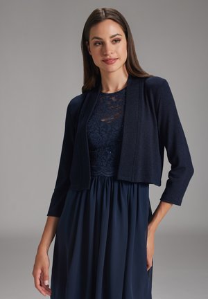 Cardigan court bleu marine à manches trois-quarts, en tissu texturé avec un léger éclat, porté sur une robe en dentelle.
