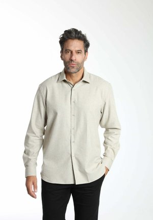 Homme aux cheveux et barbe foncés portant une chemise beige clair à boutons et un pantalon noir, debout avec une main dans la poche sur un fond blanc.