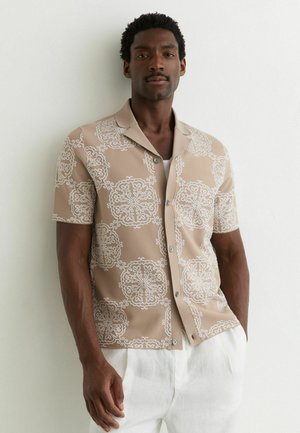 Muž nosiaci béžovú krátku košeľu na gombíky s bielymi kvetinovými vzormi a biele nohavice, stojaci oproti svetlej stene.
