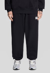 Czarne dresy z elastycznymi mankietami, z małą białą naszywką z logo. Połączone z białymi i srebrnymi butami sportowymi, na neutralnym tle.