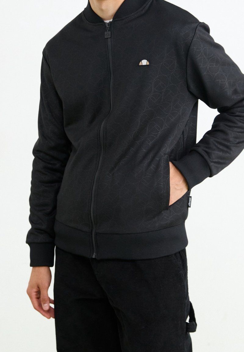 Black zip-up jacket made from textured material, featuring a subtle patterned design, ribbed cuffs, and side pockets. Small logo on chest.