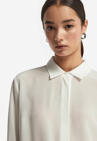Jonge vrouw met donker haar in een staart, draagt zilveren ringoorbellen en een witte blouse met kraag, kijkt recht in de camera.